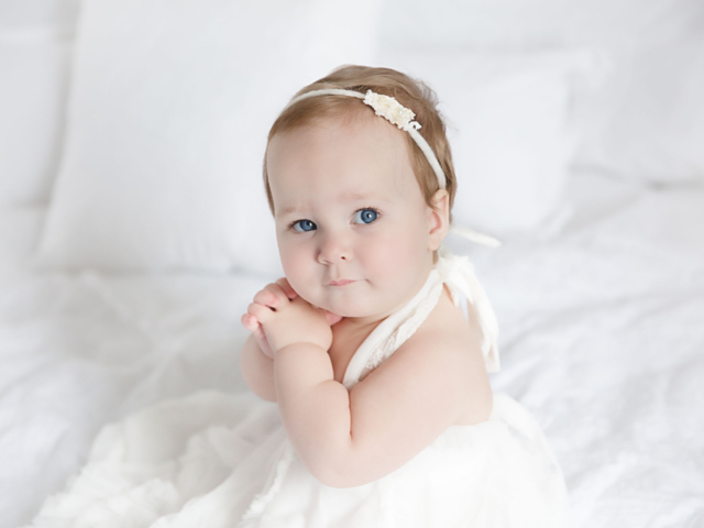 baby girl in white dress sitting on white bed during her one year portrait session by Rhonda Cunningham Photography, Lexington Ky baby photographer