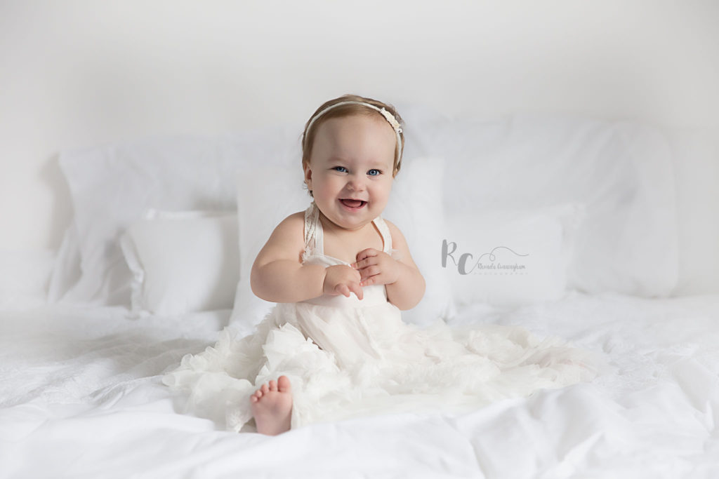 Picture of portrait part of a Lexington, KY cake smash session. One year old in white dress on white bed.